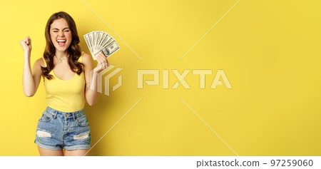 Enthusiastic smiling woman showing money cash, laughing and looking excited, standing in summer clothes against yellow background Enthusiastic smiling woman showing money cash, laughing and looking excited, standing in summer clothes against yellow background 97259060