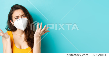Corona, pandemic and healthcare concept. Young disappointed woman complaining, shrugging and frowning upset, wearing respirator mask, standing over blue background Corona, pandemic and healthcare concept. Young disappointed woman complaining, shrugging and frowning upset, wearing respirator mask, standing over blue background 97259061