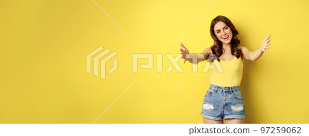 Beautiful girl in tank top, reaching hands, stretching arms forward and smiling, welcome, hugging or greeting someone, standing over yellow background 97259062