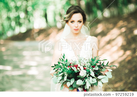 Beautiful bride in elegant white dress holding bouquet posing in park 97259219