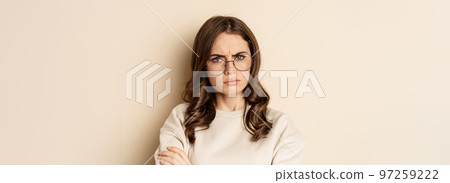 Close up portrait of complicated, troubled woman in glasses, frowning and looking displeased, standing over beige background 97259222