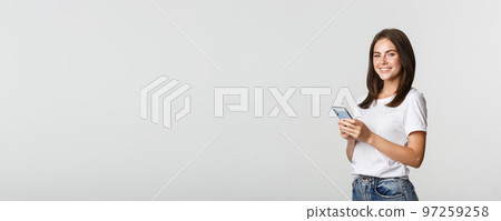 Attractive young brunette woman using mobile phone and smiling at camera Attractive young brunette woman using mobile phone and smiling at camera 97259258
