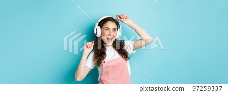 Image of cheerful young pretty girl dancing in headphones, listening music in app and jumping excited, standing over blue background Image of cheerful young pretty girl dancing in headphones, listening music in app and jumping excited, standing over blue background 97259337