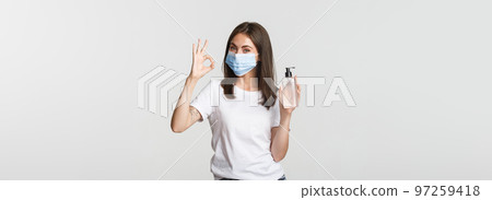 Covid-19, health and social distancing concept. Portrait of smiling brunette girl in medical mask, showing hand sanitizer and okay gesture Covid-19, health and social distancing concept. Portrait of smiling brunette girl in medical mask, showing hand sanitizer and okay gesture 97259418