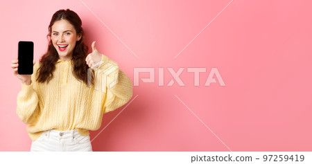 Very good app. Smiling satisfied woman showing thumbs up and empty smartphone screen, recommending app or website, standing against pink background Very good app. Smiling satisfied woman showing thumbs up and empty smartphone screen, recommending app or website, standing against pink background 97259419
