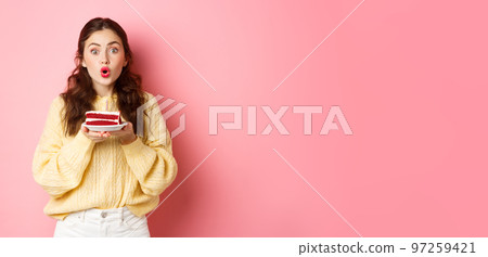 Celebration and holidays. Excited birthday girl holding b-day cake with candle and looking surprised at camera, standing against pink background Celebration and holidays. Excited birthday girl holding b-day cake with candle and looking surprised at camera, standing against pink background 97259421