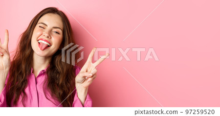 Close up of funny and happy young woman having fun, winking and showing tongue, making v-sign peace gesture, standing over pink background Close up of funny and happy young woman having fun, winking and showing tongue, making v-sign peace gesture, standing over pink background 97259520