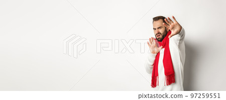 Guy asking to stop, grimacing and pulling away from camera, covering face with hands, standing over white background in red scarf and sweater Guy asking to stop, grimacing and pulling away from camera, covering face with hands, standing over white background in red scarf and sweater 97259551
