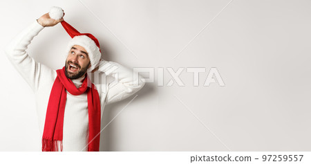 Winter holidays and New Year party concept. Happy bearded man celebrating Christmas, playing with Santa hat and looking up, white background 97259557
