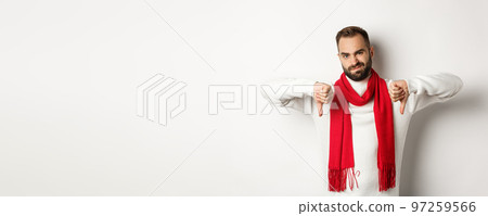 Disappointed bearded man in winter sweater and scarf, showing thumbs down and grimacing, dislike something, judging bad product, standing over white background Disappointed bearded man in winter sweater and scarf, showing thumbs down and grimacing, dislike something, judging bad product, standing over white background 97259566