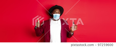 Covid-19, quarantine and holidays concept. Young african american man in medical mask, showing hand sanitizer and thumb up, recommending to use antiseptic 97259600