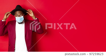 Covid-19, quarantine and holidays concept. Handsome and stylish african american man in face mask, put hat on head and looking sassy, dress-up for party, standing over red background 97259623