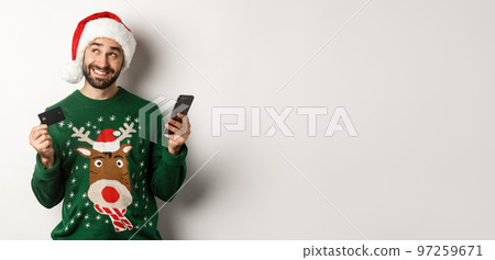 Online shopping and winter holidays concept. Smiling man thinking and holding credit card with mobile phone, wearing Santa hat, white background Online shopping and winter holidays concept. Smiling man thinking and holding credit card with mobile phone, wearing Santa hat, white background 97259671