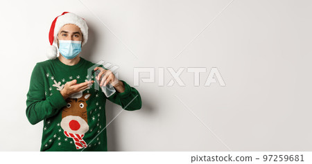 Concept of covid-19 and Christmas holidays. Caucasian man in face mask and sweater using antiseptic, clean hands with sanitizer, standing over white background Concept of covid-19 and Christmas holidays. Caucasian man in face mask and sweater using antiseptic, clean hands with sanitizer, standing over white background 97259681