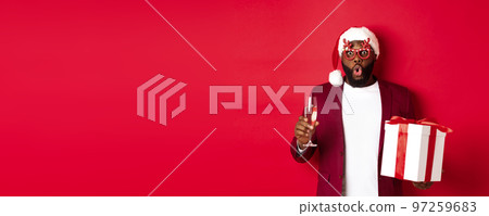 Christmas. Handsome african american man in party glasses and santa hat, holding new year gift and glass of champagne, wishing happy holidays, red background Christmas. Handsome african american man in party glasses and santa hat, holding new year gift and glass of champagne, wishing happy holidays, red background 97259683