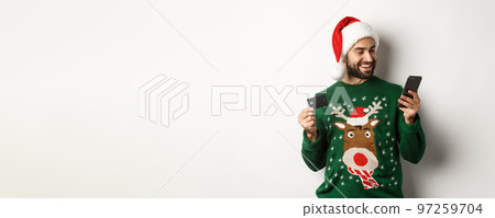 New Year, online shopping and christmas concept. Happy bearded man buying in internet with credit card and phone, wearing Santa hat and sweater, white background New Year, online shopping and christmas concept. Happy bearded man buying in internet with credit card and phone, wearing Santa hat and sweater, white background 97259704
