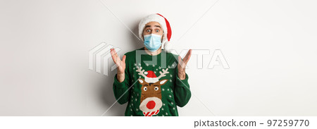 Christmas during pandemic, covid-19 concept. Surprised guy in medical mask, Santa hat and sweater celebrating New Year party, standing over white background 97259770