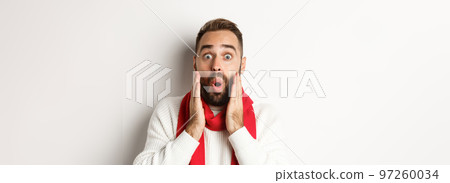 Christmas holidays. Close-up of surprised bearded guy saying wow, holding hands near face, standing against white background Christmas holidays. Close-up of surprised bearded guy saying wow, holding hands near face, standing against white background 97260034