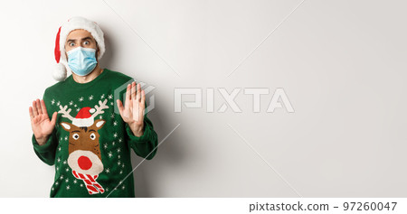 Concept of covid-19 and Christmas holidays. Anxious and freak out guy in santa hat with medical mask rejecting something, declining offer, standing over white background 97260047