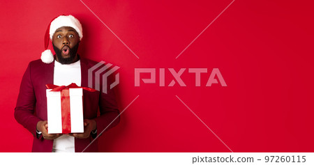 Christmas, New Year and shopping concept. Surprised african american man in santa hat saying wow, holding holiday gift and looking at camera amazed, red background 97260115