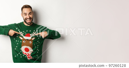 New year celebration and winter holidays concept. Cheerful man pointing at his funny Christmas sweater and smiling, white background New year celebration and winter holidays concept. Cheerful man pointing at his funny Christmas sweater and smiling, white background 97260128