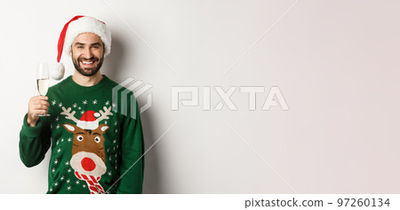 Christmas party and holidays concept. Happy man in Santa hat and sweater smiling, making a toast with glass of champagne, standing over white background 97260134