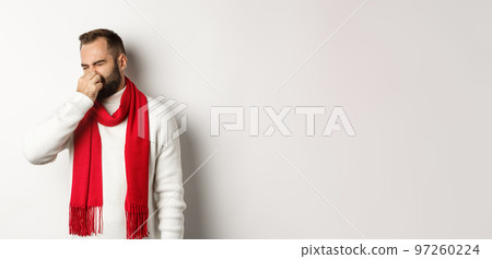 Displeased guy shut his nose from bad smell, grimacing disgusted, standing over white background in white sweater with red scarf 97260224