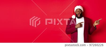Christmas, party and holidays concept. Excited Black man showing advertisement, pointing fingers right at promo offer, standing in santa hat against red background Christmas, party and holidays concept. Excited Black man showing advertisement, pointing fingers right at promo offer, standing in santa hat against red background 97260226