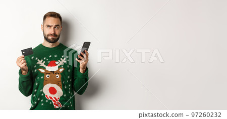 New Year, online shopping and christmas concept. Thoughtful man using mobile phone and credit card, thinking about gifts, standing over white background 97260232