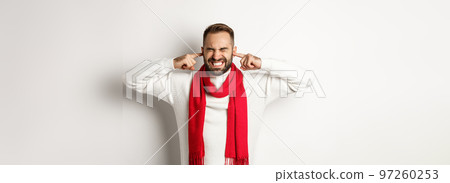 Winter holidays and shopping concept. Annoyed man shut his ears with fingers, distressed by loud noise or neighbours, standing against white background Winter holidays and shopping concept. Annoyed man shut his ears with fingers, distressed by loud noise or neighbours, standing against white background 97260253