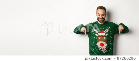 Winter holidays and christmas. Happy caucasian man in green sweater, pointing fingers down and looking satisfied, standing over white background Winter holidays and christmas. Happy caucasian man in green sweater, pointing fingers down and looking satisfied, standing over white background 97260290