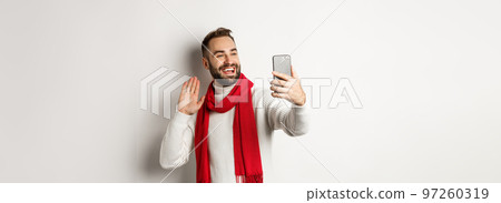 Handsome bearded man video calling friends, saying hello and waving hand at mobile phone, standing in sweater and red scarf against white background 97260319