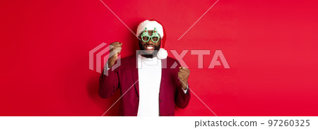 Merry Christmas. Cheerful Black man wearing funny party glasses and santa hat, smiling joyful, celebrating winter holidays, standing over red background 97260325