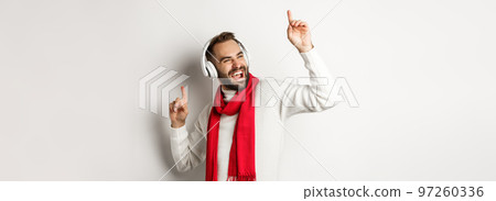 Winter holidays and technology concept. Happy man dancing to music in headphones, standing over white background in sweater Winter holidays and technology concept. Happy man dancing to music in headphones, standing over white background in sweater 97260336