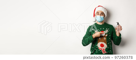 Concept of covid-19 and Christmas holidays. Young man in medical mask offer an antiseptic, pointing at hand sanitizer, wearing Santa hat, white background 97260378