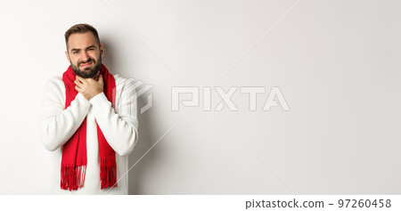 Winter, covid-19 and health concept. Sick man complaining on sore throat, touching neck and grimacing from pain, standing in sweater and red scarf, white background Winter, covid-19 and health concept. Sick man complaining on sore throat, touching neck and grimacing from pain, standing in sweater and red scarf, white background 97260458
