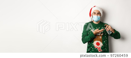 Concept of covid-19 and Christmas holidays. Caucasian man in face mask and sweater using antiseptic, clean hands with sanitizer, standing over white background 97260459