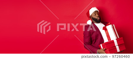 Christmas, New Year and shopping concept. Funny african american man in santa hat carry heavy xmas presents, holding gifts, standing over red background 97260460