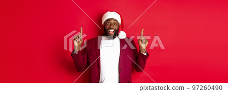Christmas, party and holidays concept. Excited Black man in santa hat and blazer, pointing fingers up and smiling, showing promo offer, red background 97260490