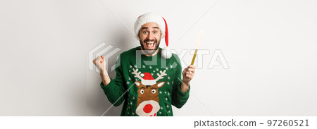 Christmas party and holidays concept. Handsome bearded man rejoicing, holding party sparkler firework and wearing Santa hat, celebrating New Year, white background Christmas party and holidays concept. Handsome bearded man rejoicing, holding party sparkler firework and wearing Santa hat, celebrating New Year, white background 97260521
