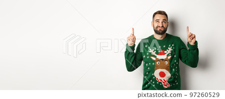 Winter holidays and christmas. Displeased guy looking doubtful at copy space, pointing fingers up and grimacing, standing over white background Winter holidays and christmas. Displeased guy looking doubtful at copy space, pointing fingers up and grimacing, standing over white background 97260529