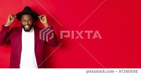 Fashion and party concept. Handsome african american man wearing hat and jacket, smiling self-assured, standing against red background Fashion and party concept. Handsome african american man wearing hat and jacket, smiling self-assured, standing against red background 97260588