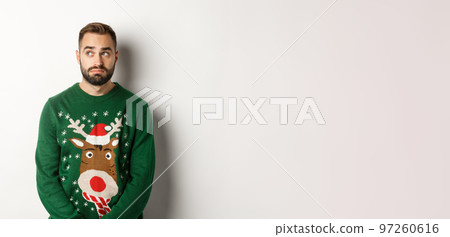 New Year, holidays and celebration. Gloomy bearded man in sweater, looking indecisive and looking aside, standing against white background 97260616