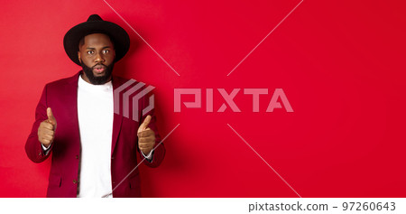 Impressed Black man showing thumbs up and staring amazed at camera, approve and praise something good, red background 97260643