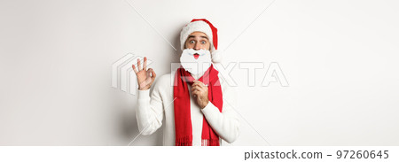 Christmas party and celebration concept. Happy male model in Santa Claus hat and white beard mask, showing ok gesture, standing over white background 97260645