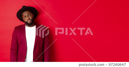 Sad and unamused Black man looking with pity at camera, frowning disappointed, standing against red background 97260647