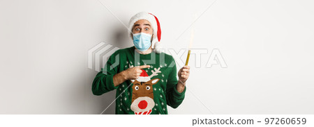 Concept of covid-19 and Christmas holidays. Excited young man in face mask, pointing finger at party sparkler, celebrating New Year during quarantine, standing over white background 97260659