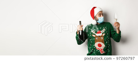 Concept of covid-19 and Christmas holidays. Man in medical mask and Santa hat enjoying glass of champagne, celebrating New Year, standing over white background Concept of covid-19 and Christmas holidays. Man in medical mask and Santa hat enjoying glass of champagne, celebrating New Year, standing over white background 97260797