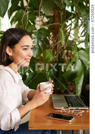 Vertical shot of beautiful woman sits in cafe with laptop, works or studies online at co-working space, smiling relaxed Vertical shot of beautiful woman sits in cafe with laptop, works or studies online at co-working space, smiling relaxed 97260826