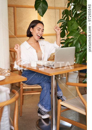 Enthusiastic asian girl celebrating, looking at laptop and triumphing, makes fist pumps with excited face, sits in cafe Enthusiastic asian girl celebrating, looking at laptop and triumphing, makes fist pumps with excited face, sits in cafe 97260848